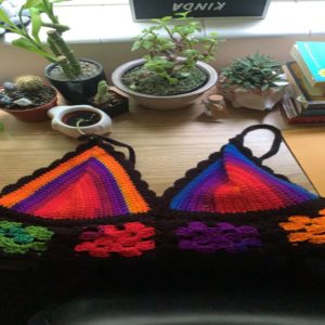 A custom crochet colorful bikini top lays on top of a wooden desk in front of little plants.