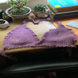 A custom crochet lavender purple bikini top lays on top of a wooden desk in front of little plants and books.
