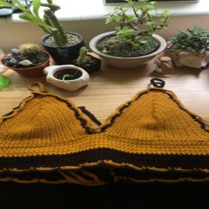 A custom crochet mustard yellow bikini top lays on top of a wooden desk in front of little plants.